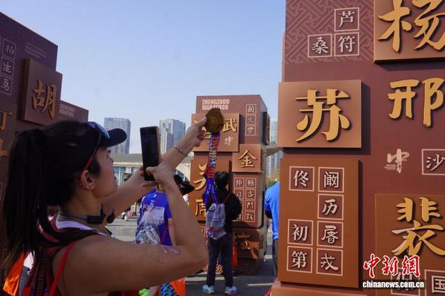 11月16日，河南郑州，一名参赛选手完赛后举起奖牌在姓氏墙前拍照。韩章云 摄